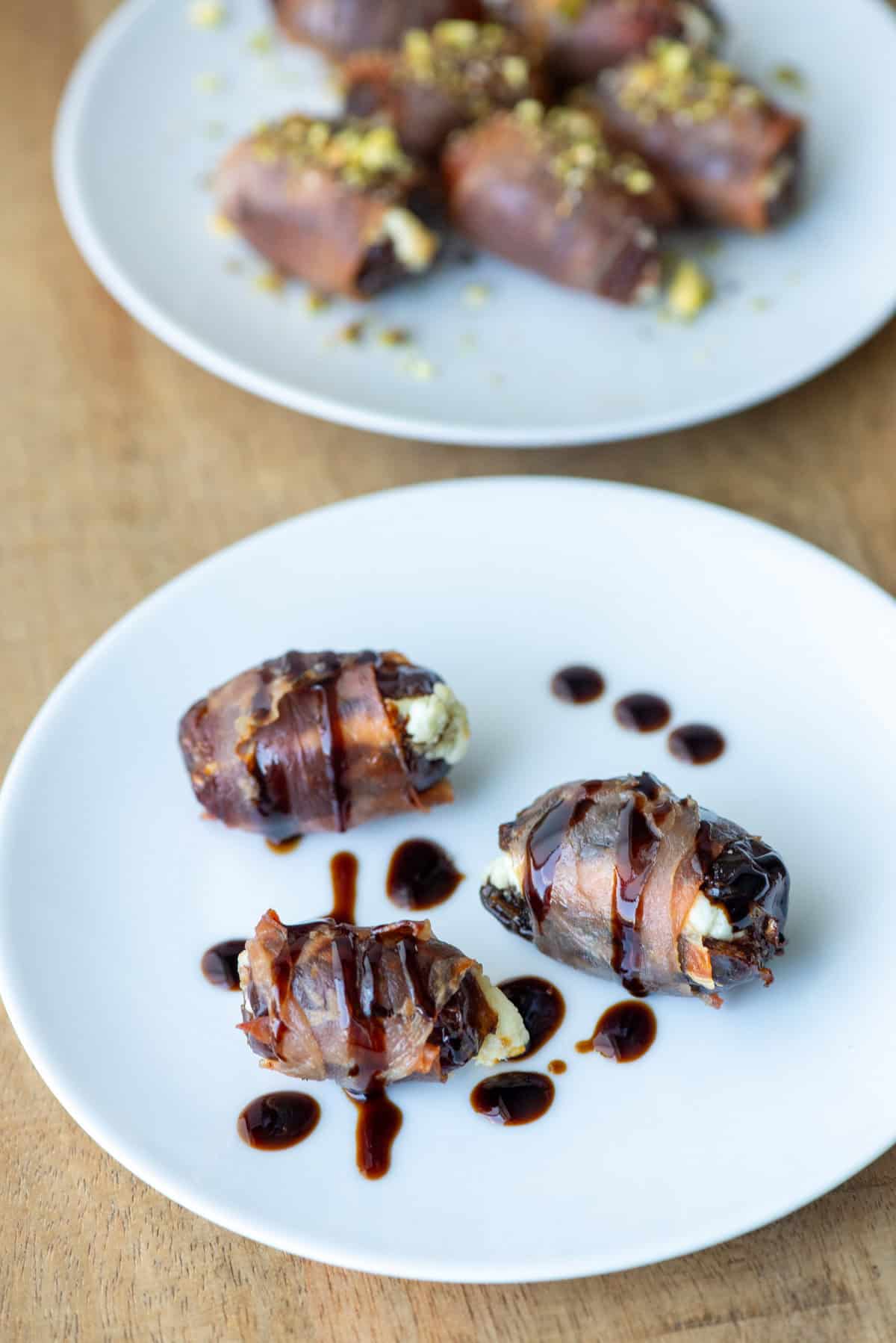 Three prosciutto wrapped dates with balsamic glaze drizzle in front of a plate with other prosciutto wrapped dates topped with hot honey and chopped pistachios