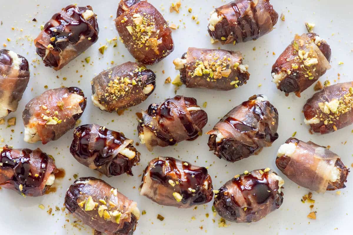 Overhead view of randomly placed prosciutto wrapped dates on a white plate, some topped with balsamic glaze and some topped with hot honey and chopped pistachios