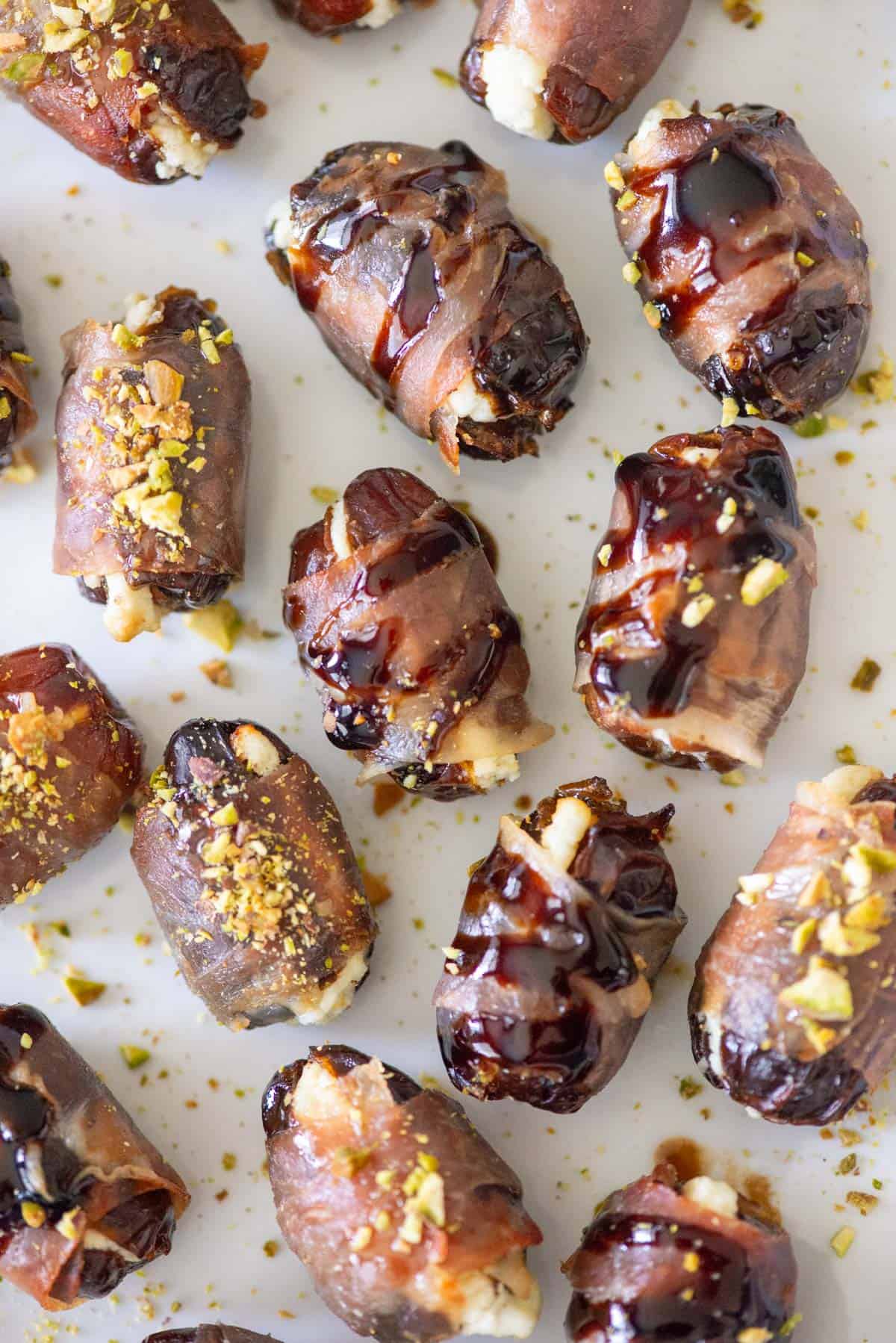 Overhead view of randomly placed prosciutto wrapped dates on a white plate, some topped with balsamic glaze and some topped with hot honey and chopped pistachios