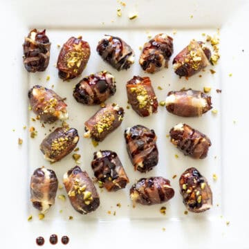 Overhead view of randomly placed prosciutto wrapped dates on a white serving platter, some topped with balsamic glaze and some topped with hot honey and chopped pistachios