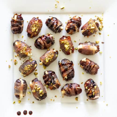 Overhead view of randomly placed prosciutto wrapped dates on a white serving platter, some topped with balsamic glaze and some topped with hot honey and chopped pistachios