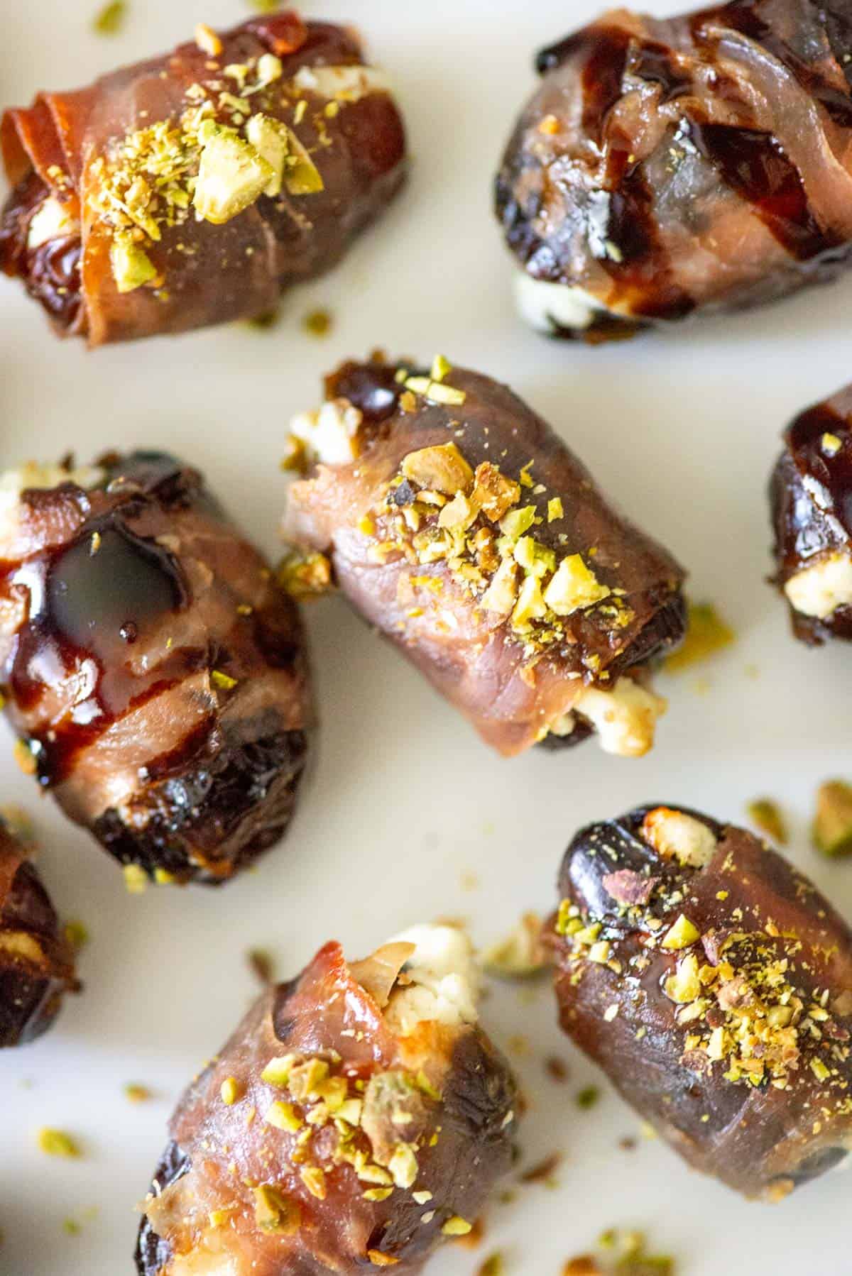 Close up of randomly placed prosciutto wrapped dates on a white plate, some topped with balsamic glaze and some topped with hot honey and chopped pistachios