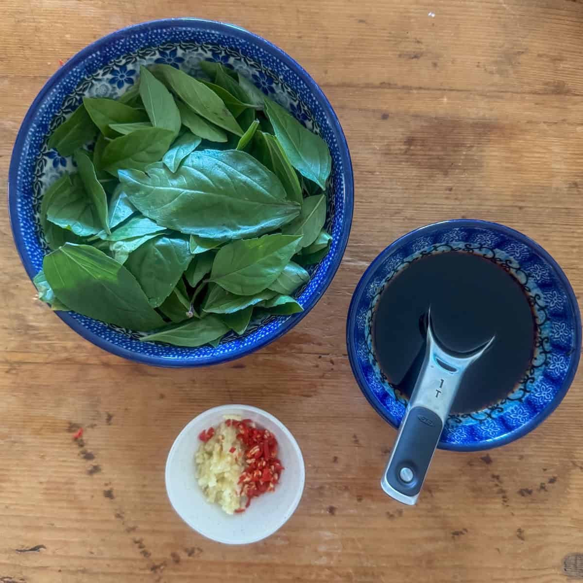 Overhead photo of Thai basil leaves in a bowl, the stir fry sauce mixed and a small bowl with garlic and Thai red chilies