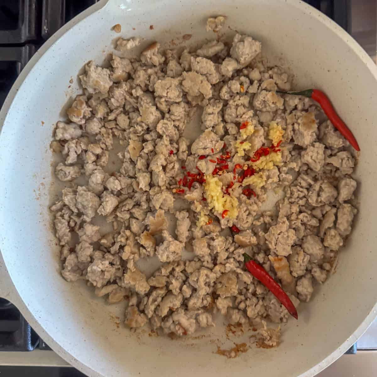 Ground chicken that's cooking in a skillet with raw garlic, chopped Thai red chilies and two whole Thai red chilies