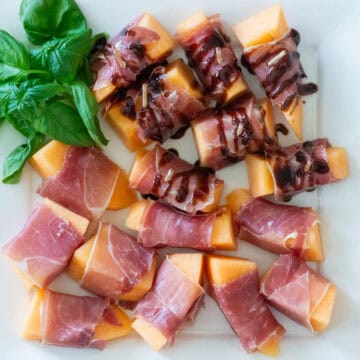 Overhead view of prosciutto wrapped melon slices on a white platter with basil garnish in the corner, half of them have a balsamic drizzle