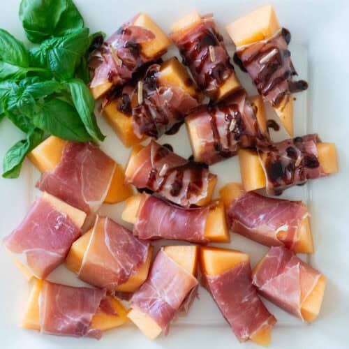 Overhead view of prosciutto wrapped melon slices on a white platter with basil garnish in the corner, half of them have a balsamic drizzle