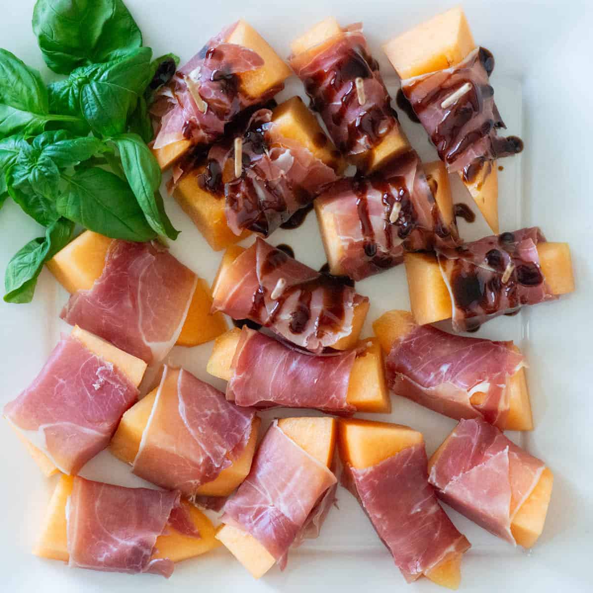 Overhead view of prosciutto wrapped melon slices on a white platter with basil garnish in the corner, half of them have a balsamic drizzle