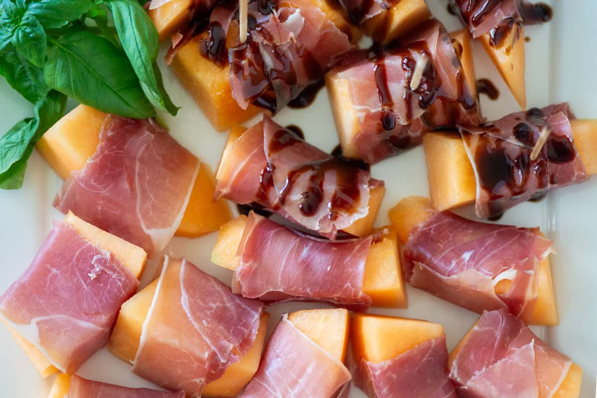 Overhead close up view of prosciutto wrapped cantaloupe slices on a white platter with basil garnish in the corner, half of them have a balsamic drizzle