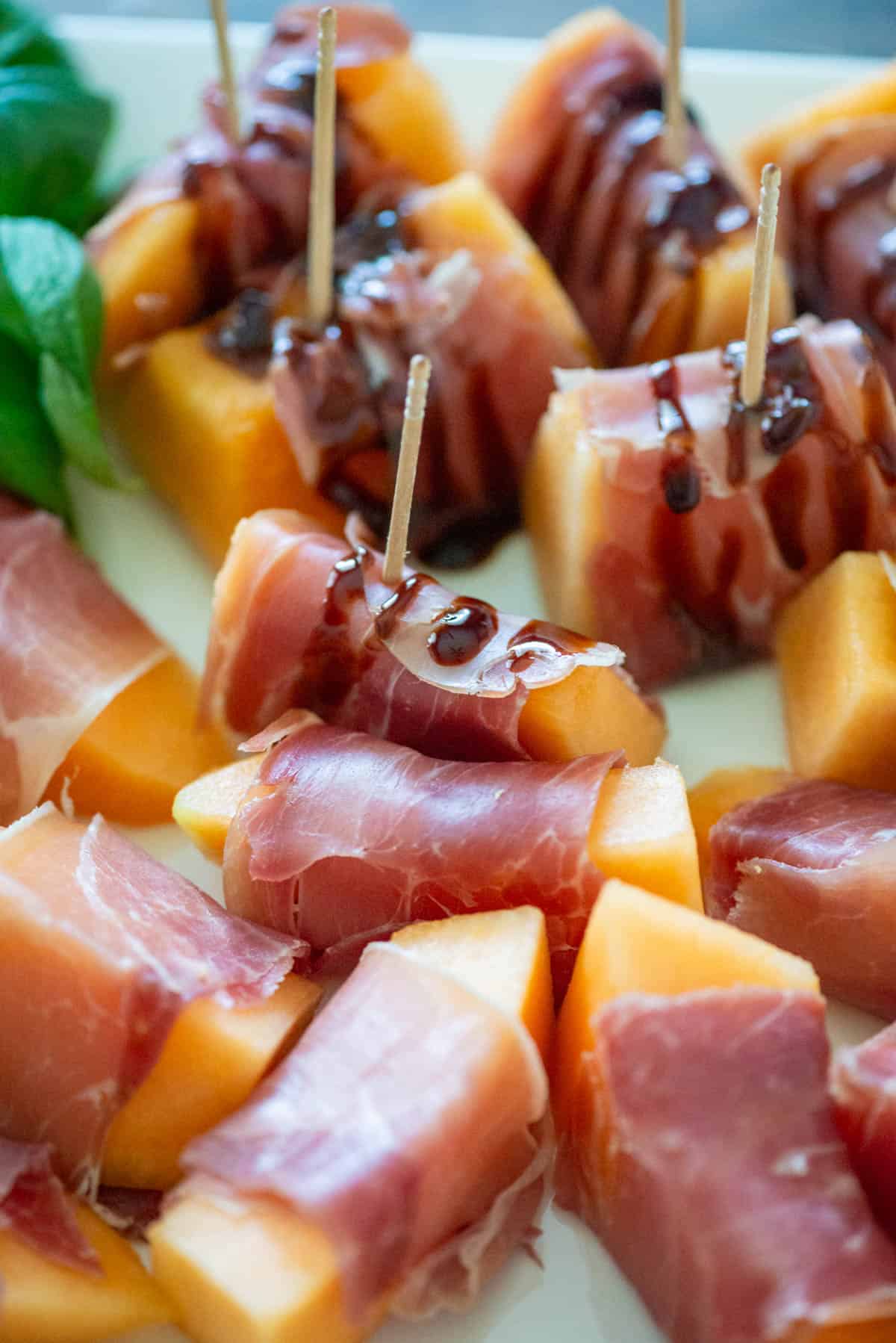 Overhead view of prosciutto wrapped melon slices on a white platter, half of them have a balsamic drizzle and a toothpick sticking out of the top