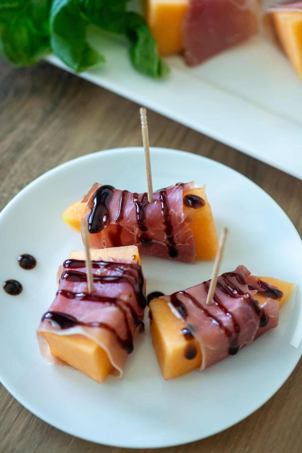 Overhead view of three pieces of prosciutto wrapped melon small plate drizzled with balsamic glaze on a small plate with a platter of prosciutto wrapped melon with basil in the background