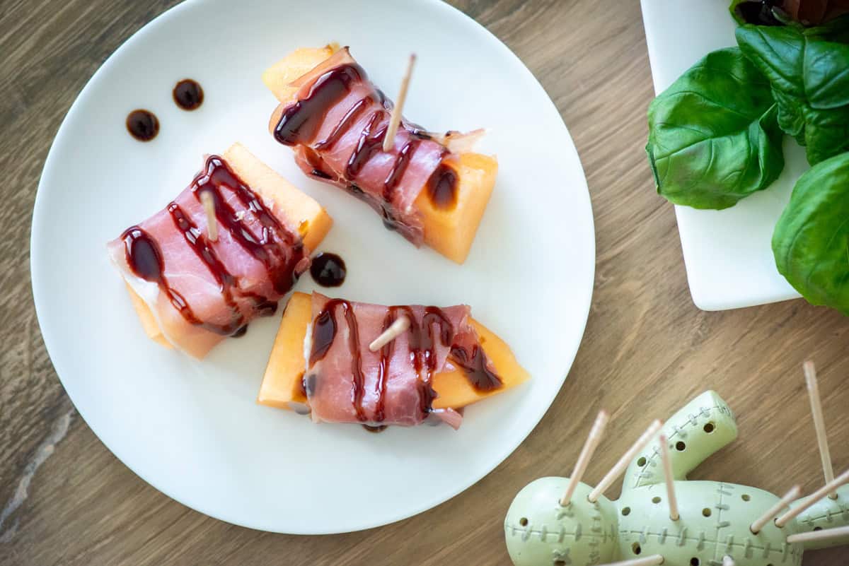 Overhead view of three pieces of prosciutto wrapped melon small plate drizzled with balsamic glaze on a small plate with basil and and a toothpick holder on the side
