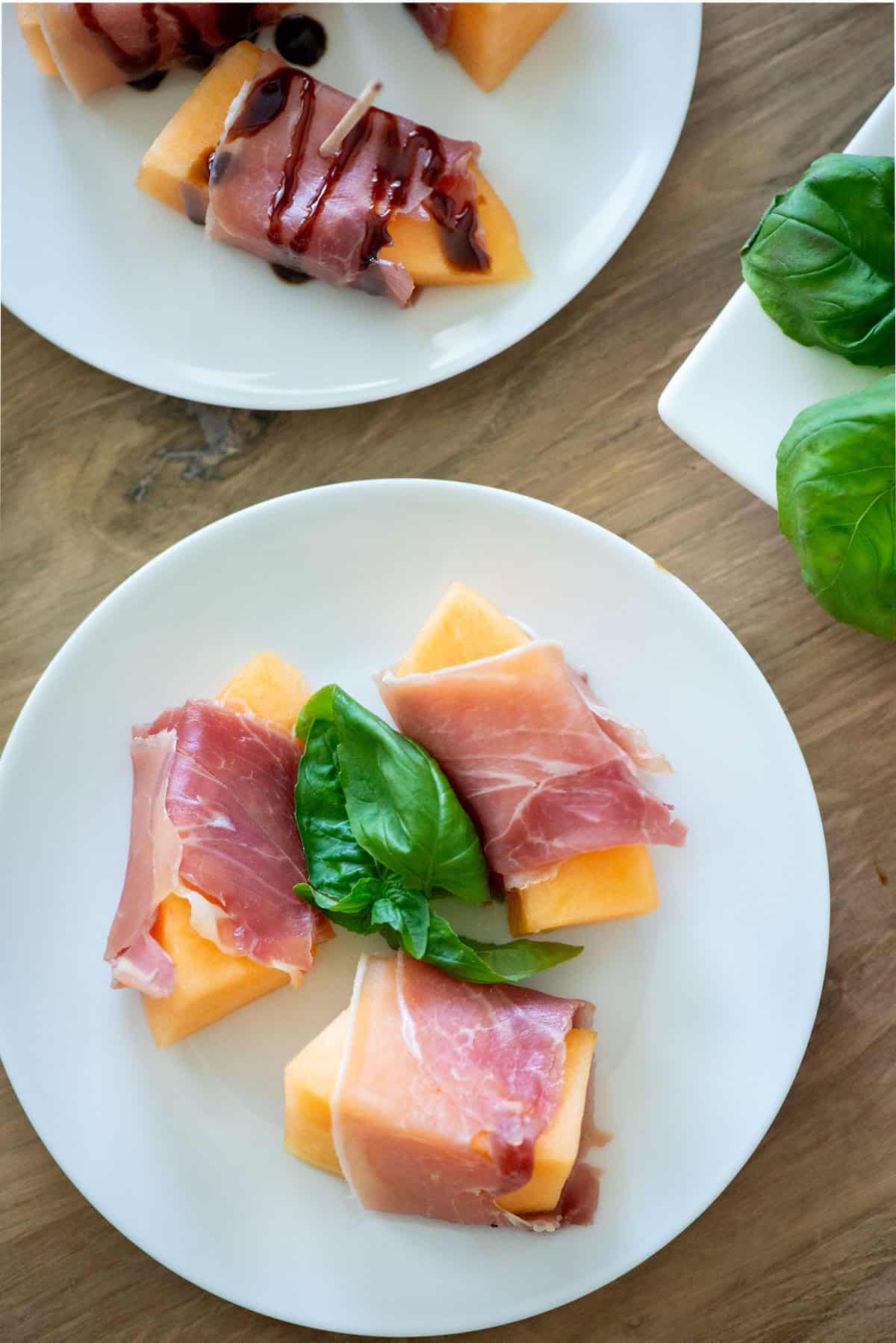 Overhead view of three pieces of prosciutto wrapped melon small plate drizzled and a piece of basil on a small plate with another plate of prosciutto wrapped melon that's drizzled with balsamic glaze in the background