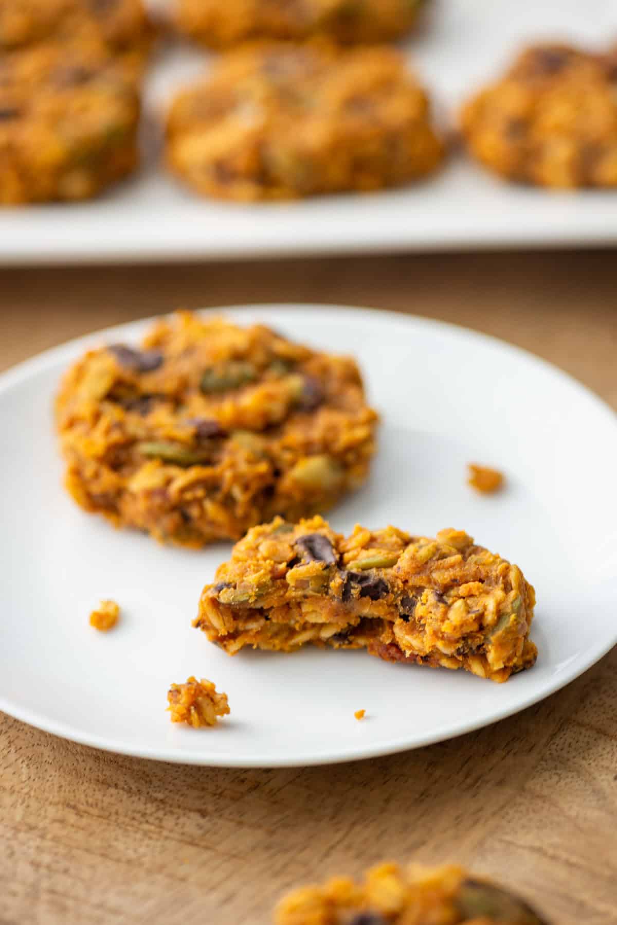 Overhead angled view of two pumpkin breakfast cookies on a white plate, one with a bite taken out, sitting in front of a platter with more cookies in the background