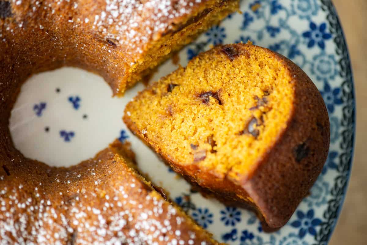 Overhead close up photo of a slice of pumpkin date bread made in a bundt pan that's been sliced and turned over on it's side on a cake platter so that you can see the inside of the bread. Pumpkin date bread that's been sprinkled with powdered sugar and has not been cut is partially visible