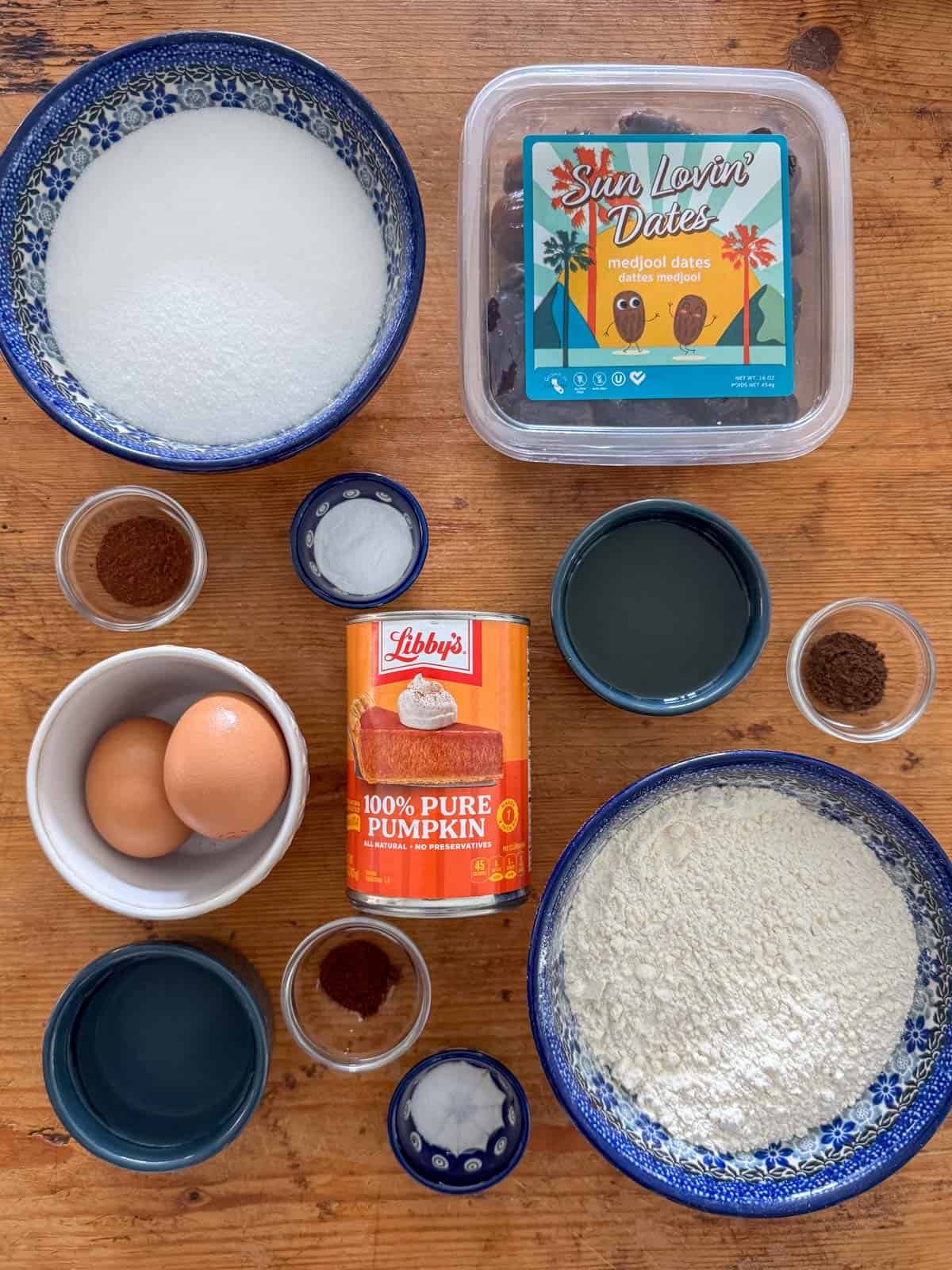 Overhead photo of ingredients need to make pumpkin date bread: flour, baking soda, salt, cinnamon, cloves, allspice, white sugar, neutral oil, eggs, canned pumpkin, water and dates.