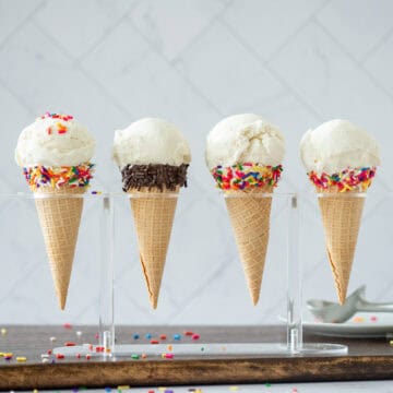 Side view of 4 homemade vanilla bean ice cream in sugar cones in an ice cream holder. 3 sugar cones have a band of multicolored sprinkles at the opening and 1 has chocolate sprinkles