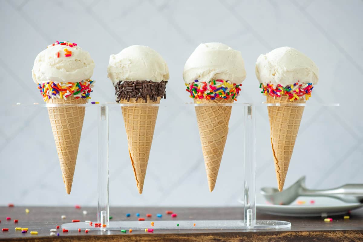 Side view of 4 homemade vanilla bean ice cream in sugar cones in an ice cream holder. 3 sugar cones have a band of multicolored sprinkles at the opening and 1 has chocolate sprinkles