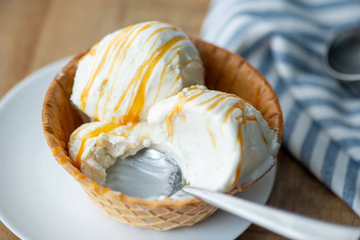 Three scoops of homemade vanilla bean ice cream drizzled with homemade salted caramel sauce in a waffle cone bowl with a spoon sticking out