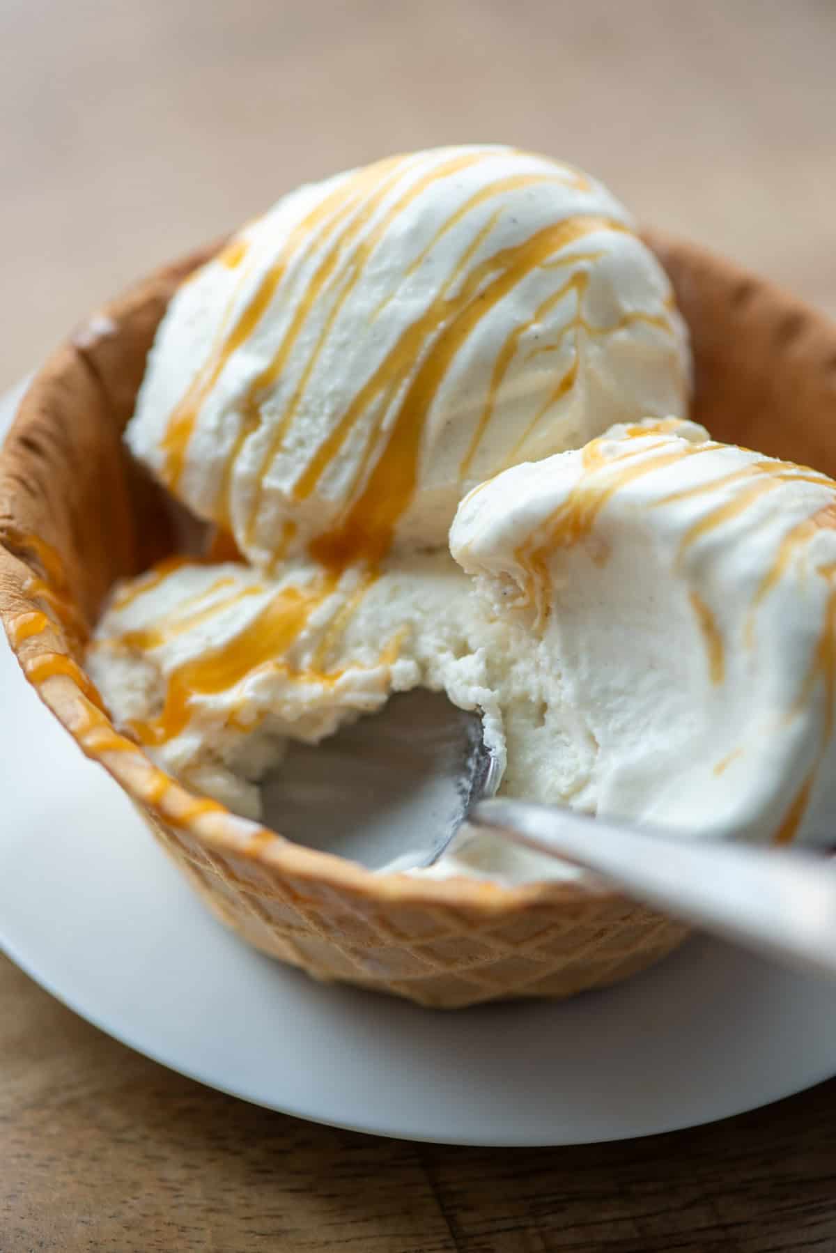 Close up view of 3 scoops of homemade vanilla bean ice cream drizzled with homemade salted caramel sauce in a waffle cone bowl with a spoon sticking out