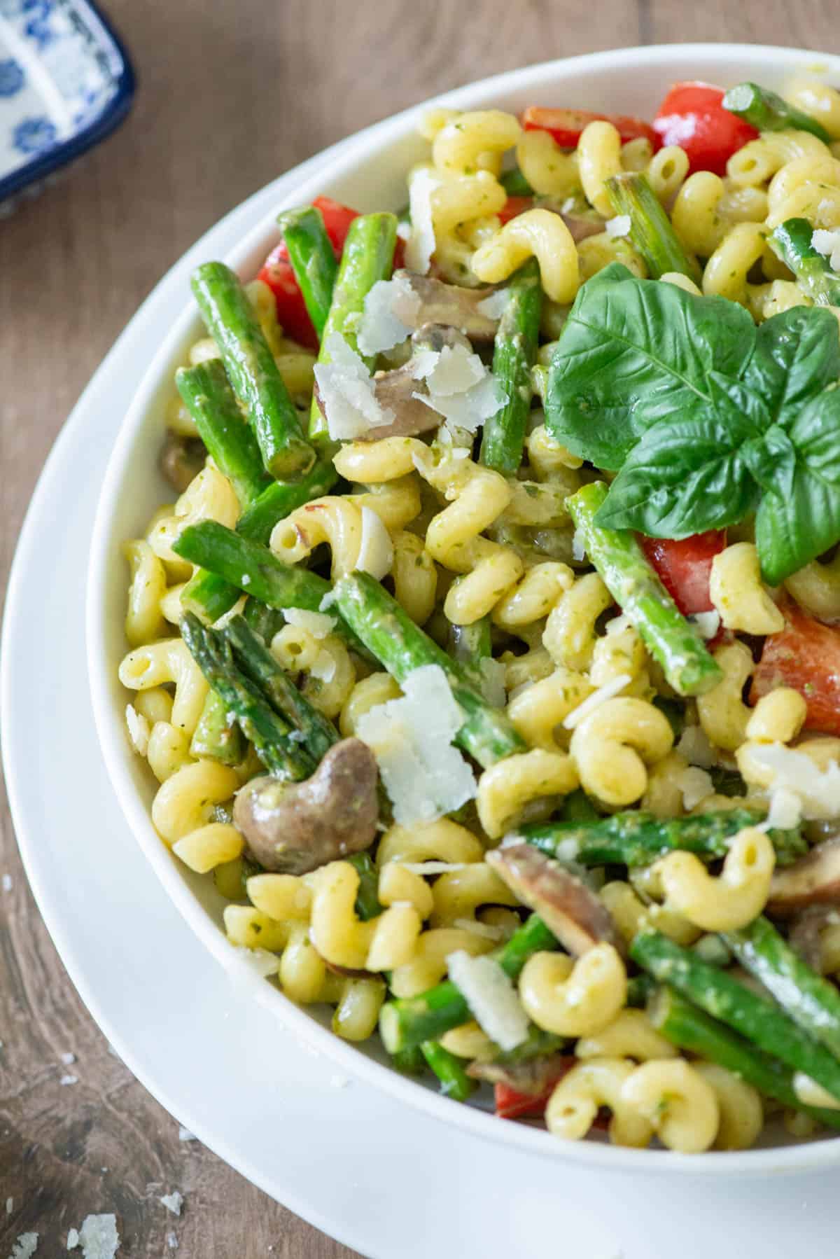 Overhead angled view of a plate of vegetable pesto pasta made with asparagus, red bell peppers and mushrooms and garnished with extra parmesan cheese and basil on top of another white plate