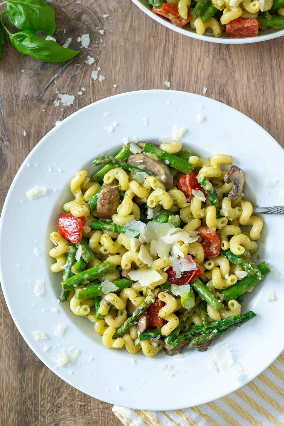 Overhead view of a white plate of vegetable pesto pasta garnished with extra parmesan cheese with a fork sticking out of the right side with another basil scattered on the table and another plate of vegetable pesto pasta partially visible in the background