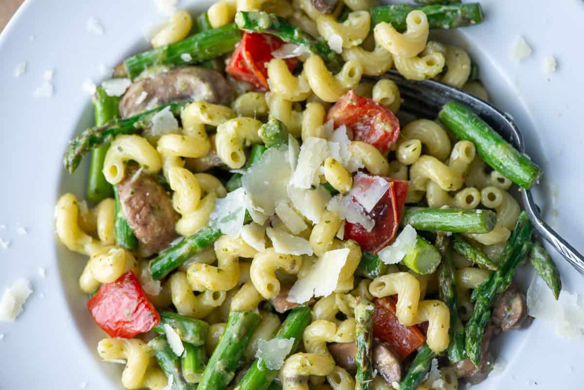 Overhead close up view of a partial white plate of vegetable pesto pasta made with asparagus, red bell peppers and mushrooms and garnished with extra parmesan cheese with a fork sticking out of the right side