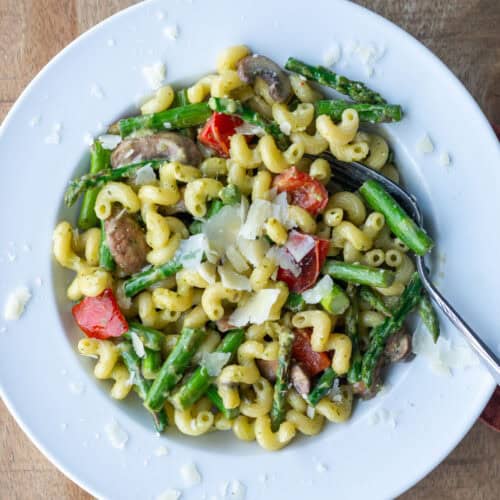 Overhead close up view of a white pasta plate of vegetable pesto pasta made with asparagus, red bell peppers and mushrooms and garnished with extra parmesan cheese with a fork sticking out of the right side