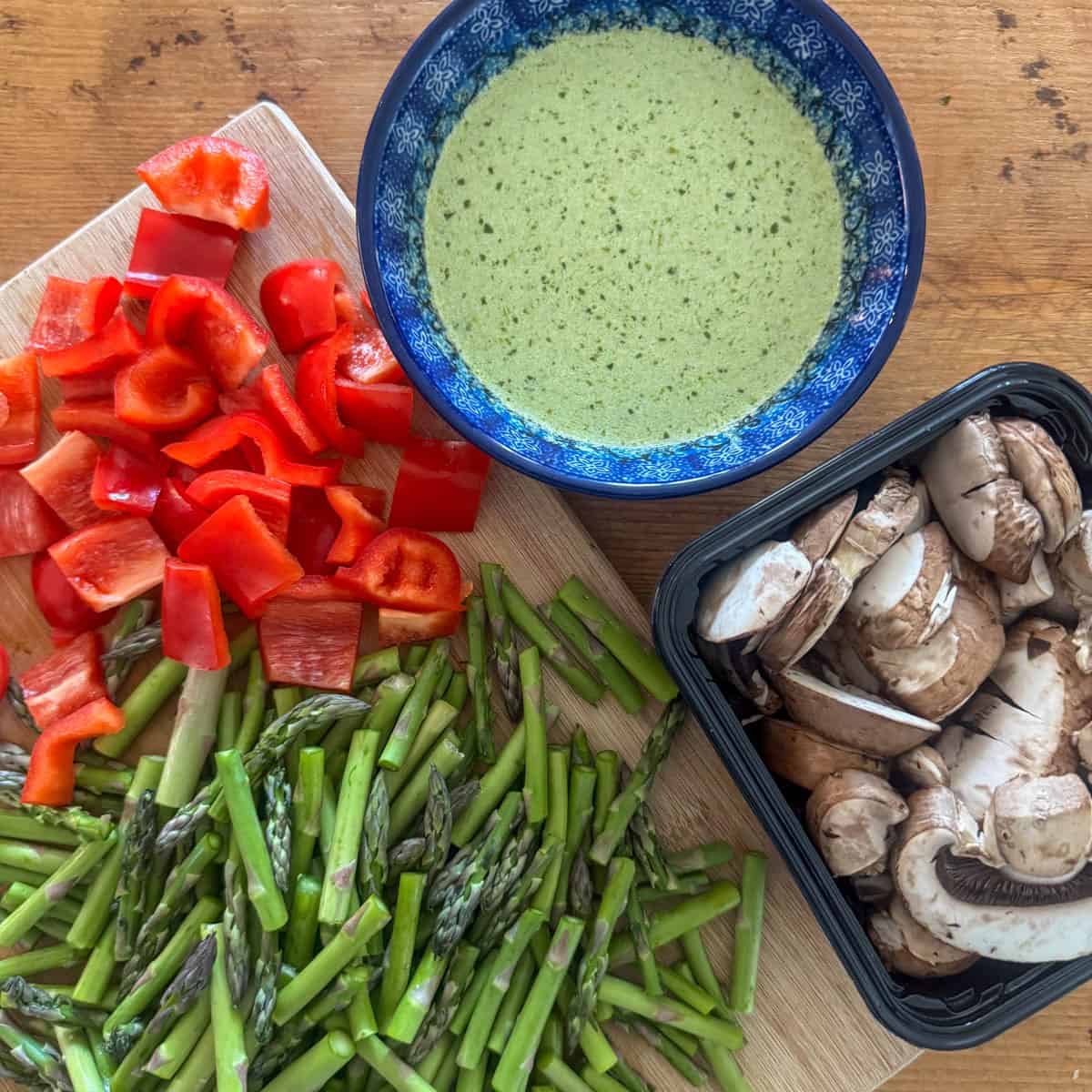 Overhead photo of cut red bell peppers, asparagus and mushrooms with a bowl of basil pesto and heavy shipping cream mixed together