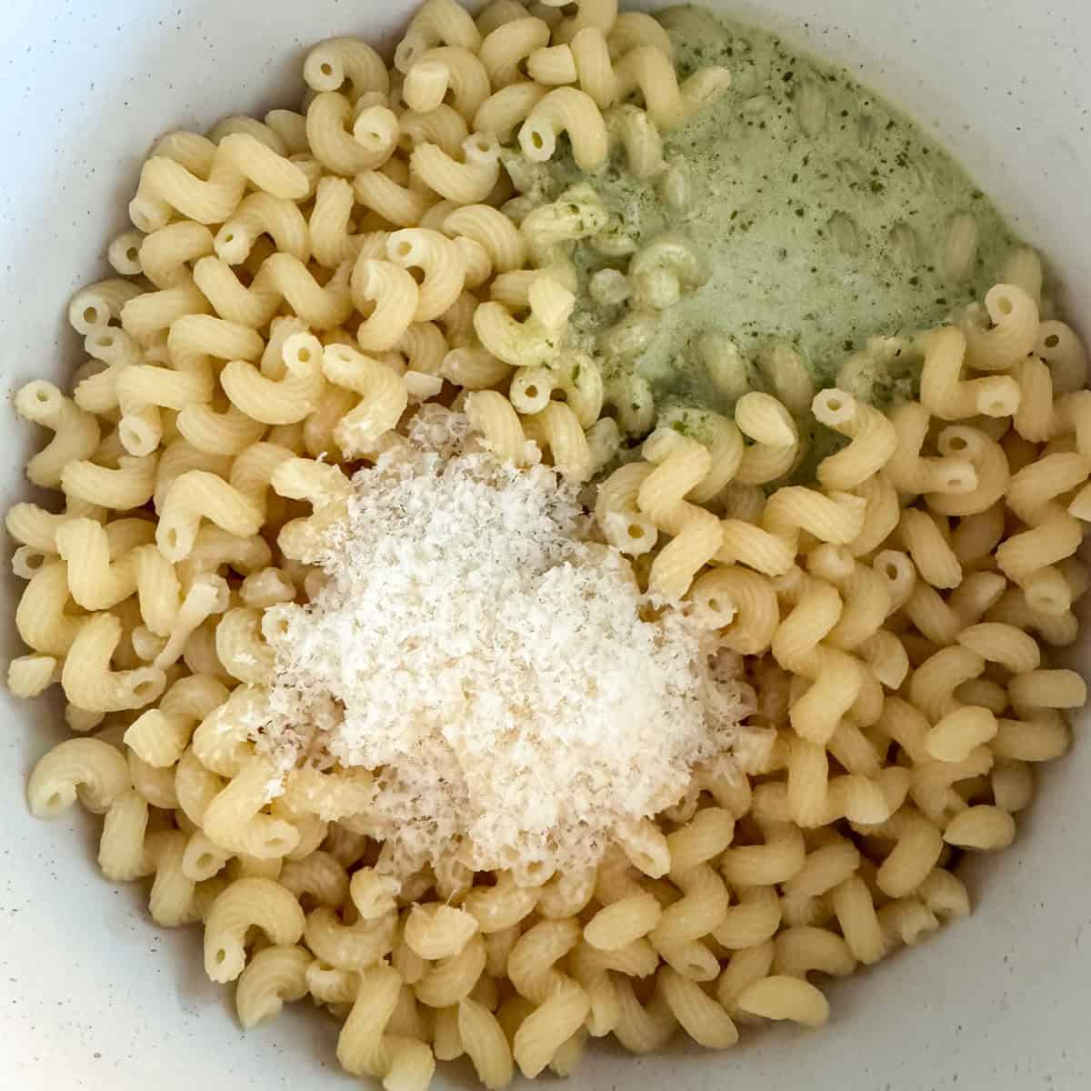 Overhead photo of cooked pasta in a pasta pot with a basil pesto and heavy cream mixture and parmesan cheese that needs to be stirred all together