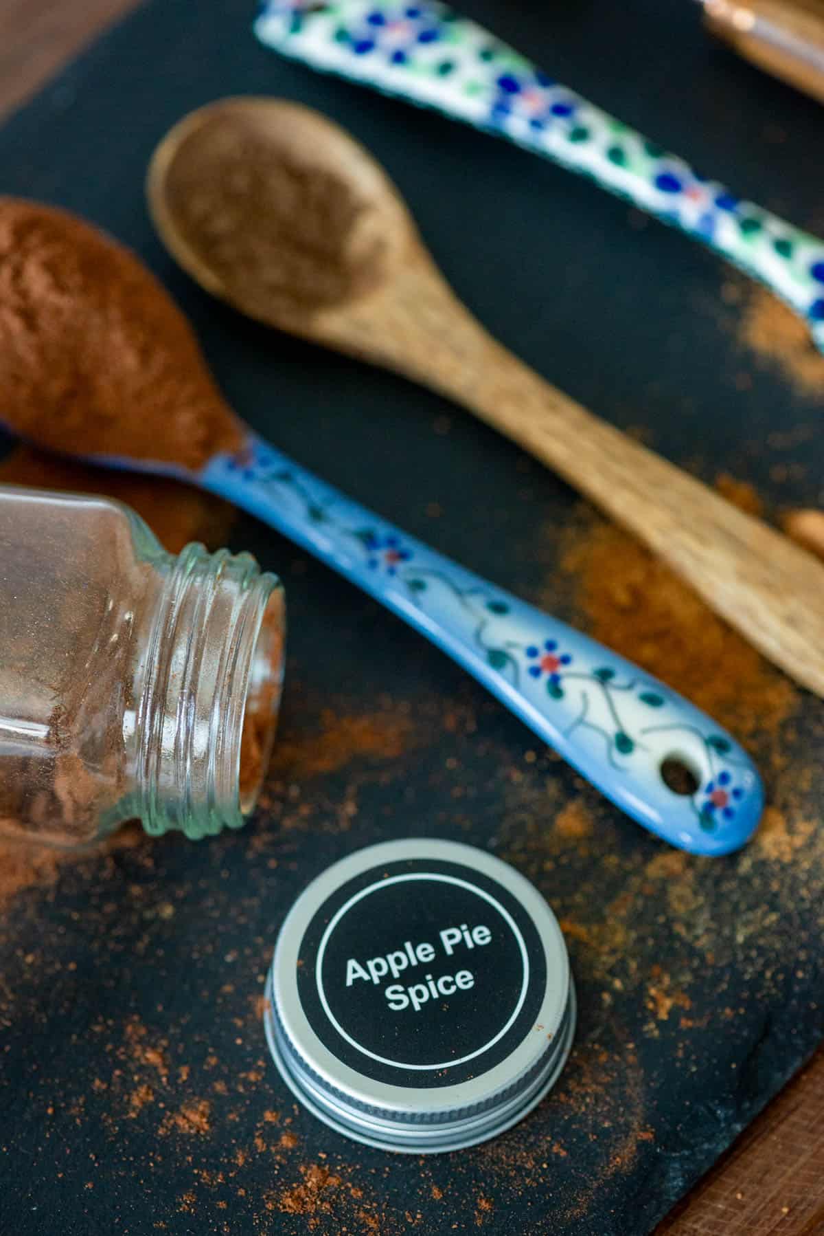Angled overhead view of 3 different types of teaspoons all with different spices on them with a spilled spice jar in front with the cap labeled Apple Pie Spice off and lying on the platter