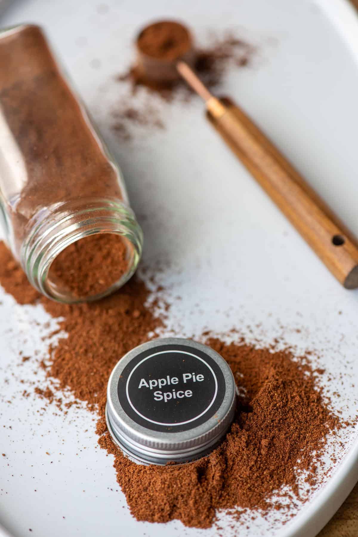 Overhead photo of a white platter with an apple pie spice jar on it's side with apple pie spice spilling out with the lid to the jar, which says Apple Pie Spice, in the middle of the loose spice and a teaspoon filled with spice in the background