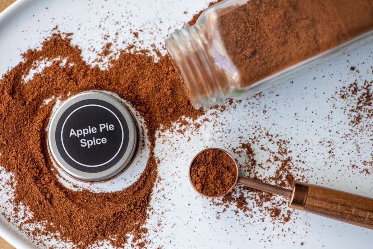 Overhead photo of a white platter with an apple pie spice jar on it's side with apple pie spice spilling out with the lid to the jar, which says Apple Pie Spice, in the middle of the loose spice and a teaspoon filled with spice to the side