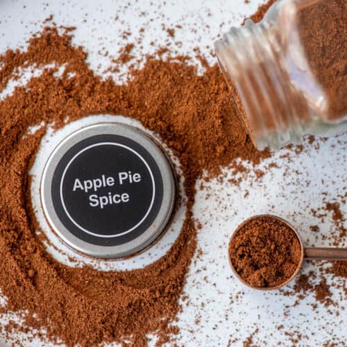 Overhead photo of an apple pie spice jar on it's side with apple pie spice spilling out with the lid to the jar, which says Apple Pie Spice, in the middle of the loose spice and a teaspoon filled with spice to the side