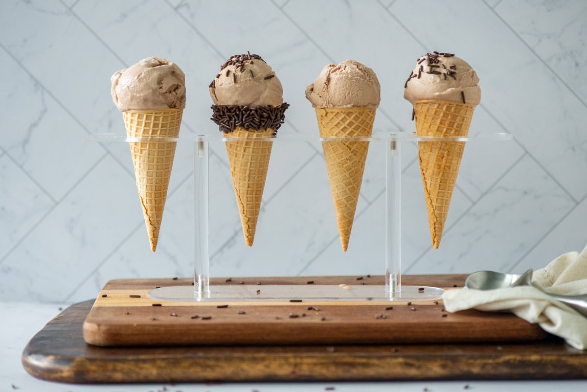 Side view of 4 homemade chocolate malt ice cream sugar cones in an ice cream holder. 1 sugar cone has a band of chocolate sprinkles at the opening and 2 have chocolate sprinkles on top