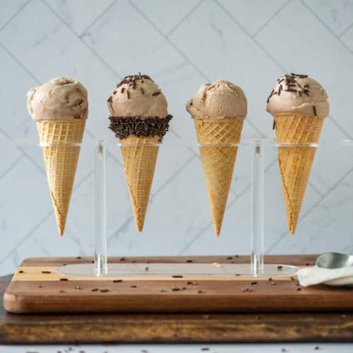 Side view of 4 homemade chocolate malt ice cream sugar cones in an ice cream holder. 1 sugar cone has a band of chocolate sprinkles at the opening and 2 have chocolate sprinkles on top