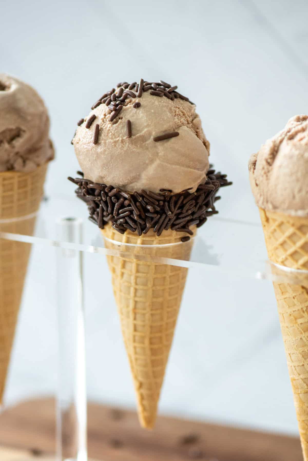 Close up photo of a chocolate malt ice cream cone in a holder with two other chocolate malt ice cream cones partially visible on the sides. The main cone also has chocolate sprinkles.
