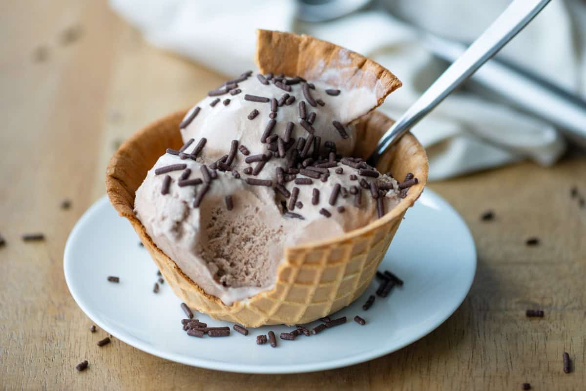 Side view photo of three scoops of chocolate malt ice cream in a waffle cone bowl with a chunk of the bowl broken off and stuck into the top scoop of ice cream. There's also a spoon sticking out of the bowl and an ice cream scoop in the background.
