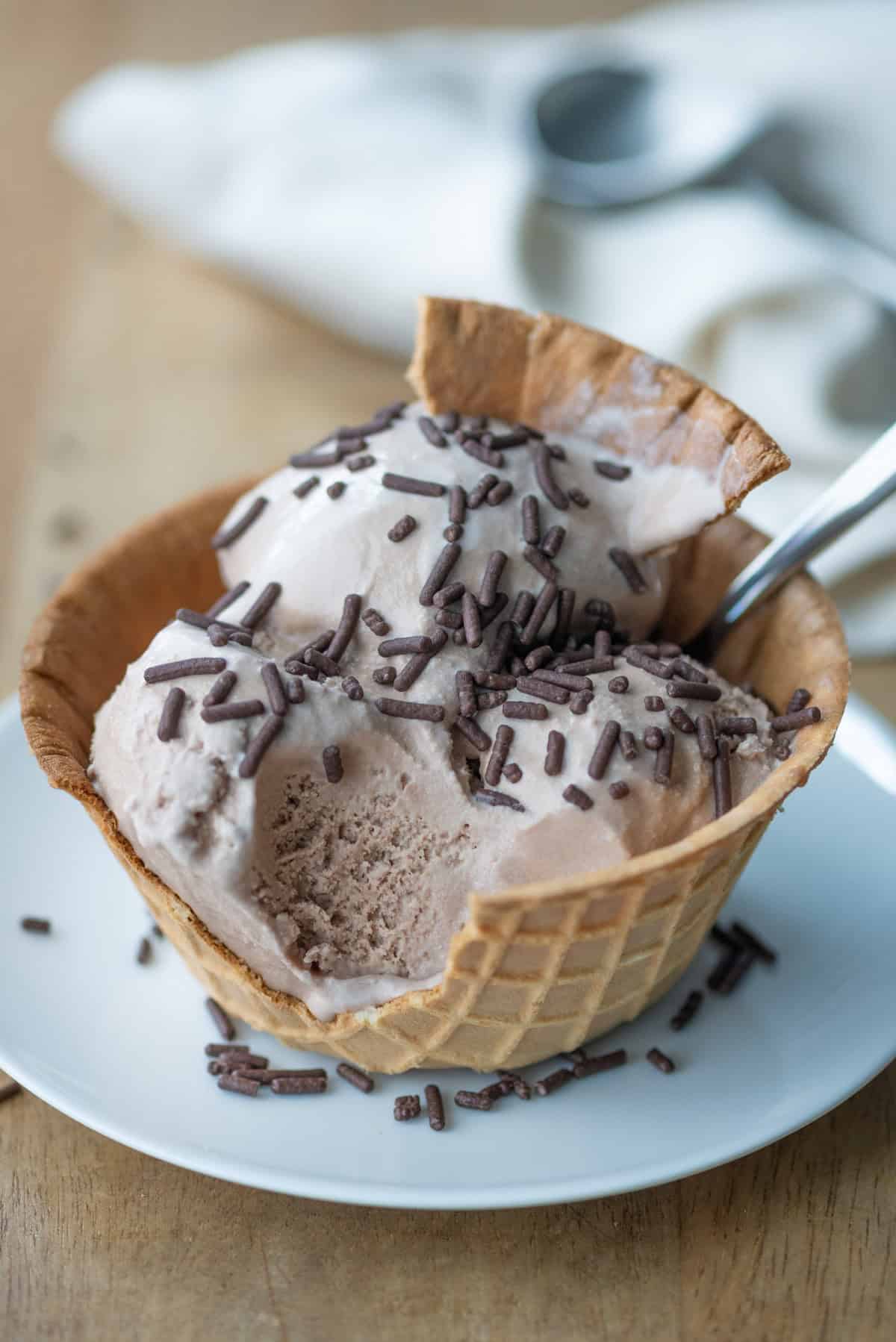 Side view photo of three scoops of chocolate malt ice cream in a waffle cone bowl with a chunk of the bowl broken off and stuck into the top scoop of ice cream. There's also a spoon sticking out of the bowl and an ice cream scoop in the background.