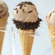 Close up photo of a chocolate malt ice cream cone in a holder with two other chocolate malt ice cream cones partially visible on the sides. The main cone also has chocolate sprinkles. It also has a title overlay at the top that says Chocolate Malt Ice Cream