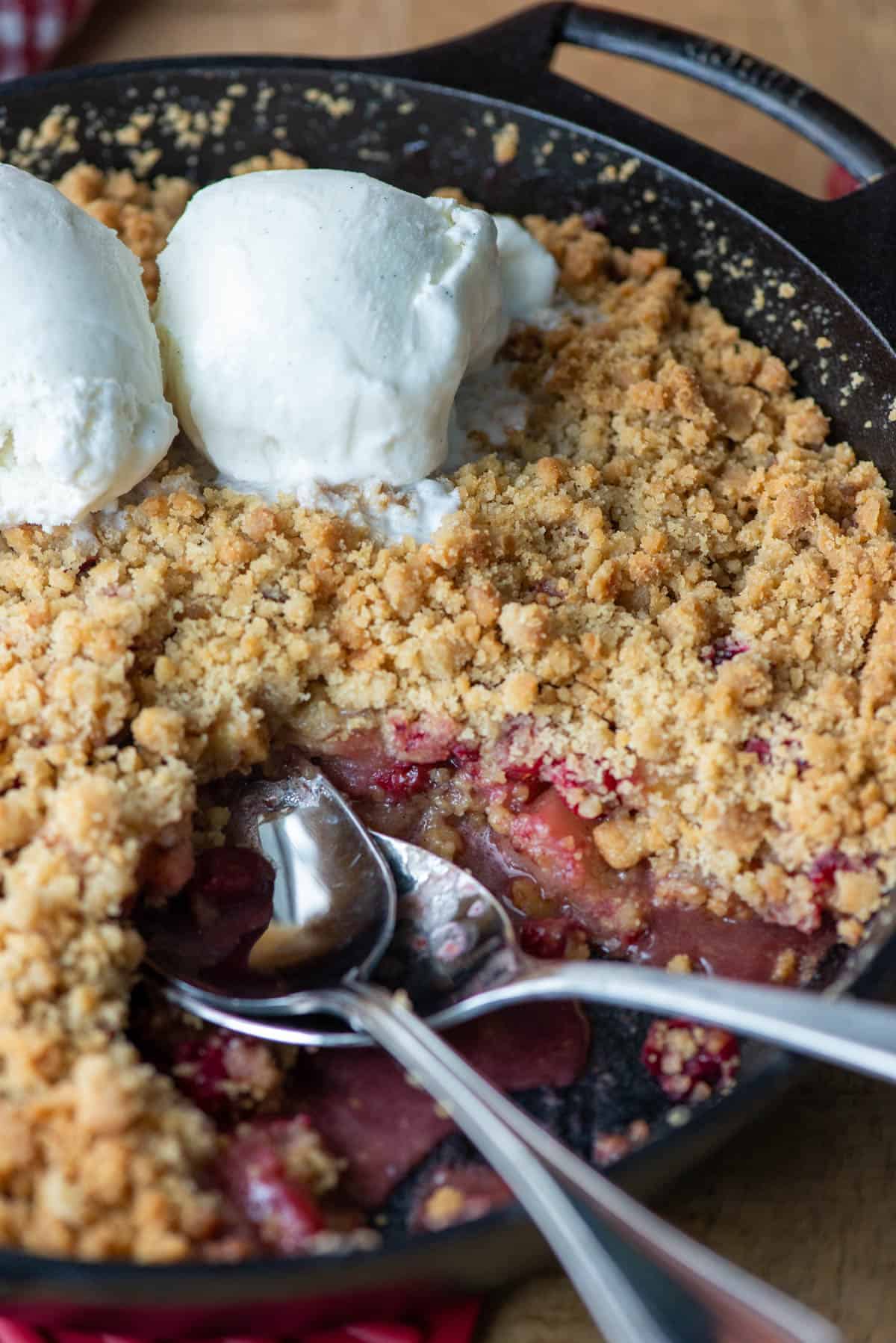 Overhead angled photo of cranberry apple crumble in a cast iron pan with a scoop of vanilla bean ice cream melting on top and two spoon sticking out