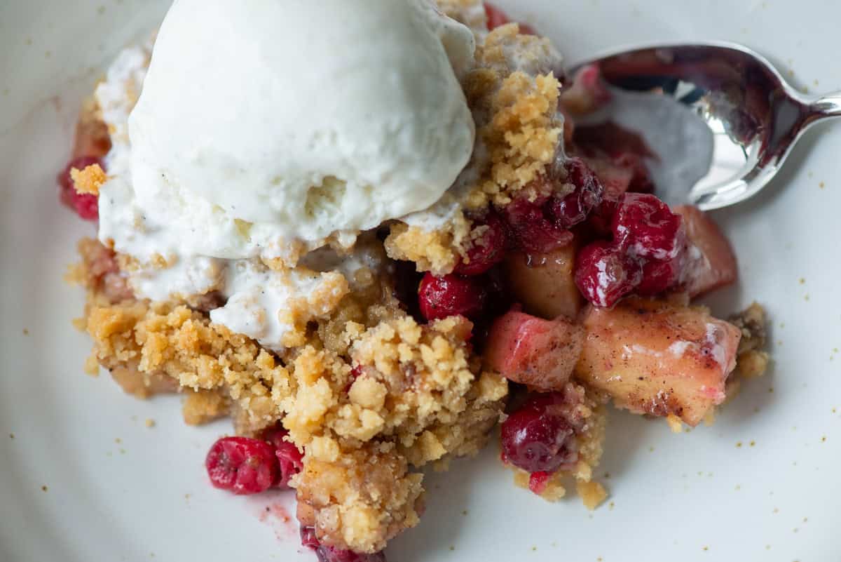 Close up of cranberry apple crumple on a plate with ice cream melting on top and a spoon sticking out of the side