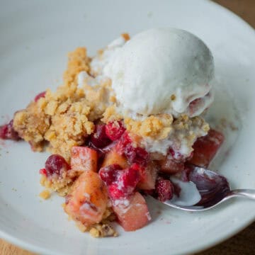 Close up of cranberry apple crumple on a plate with ice cream melting on top and a spoon sticking out of the side