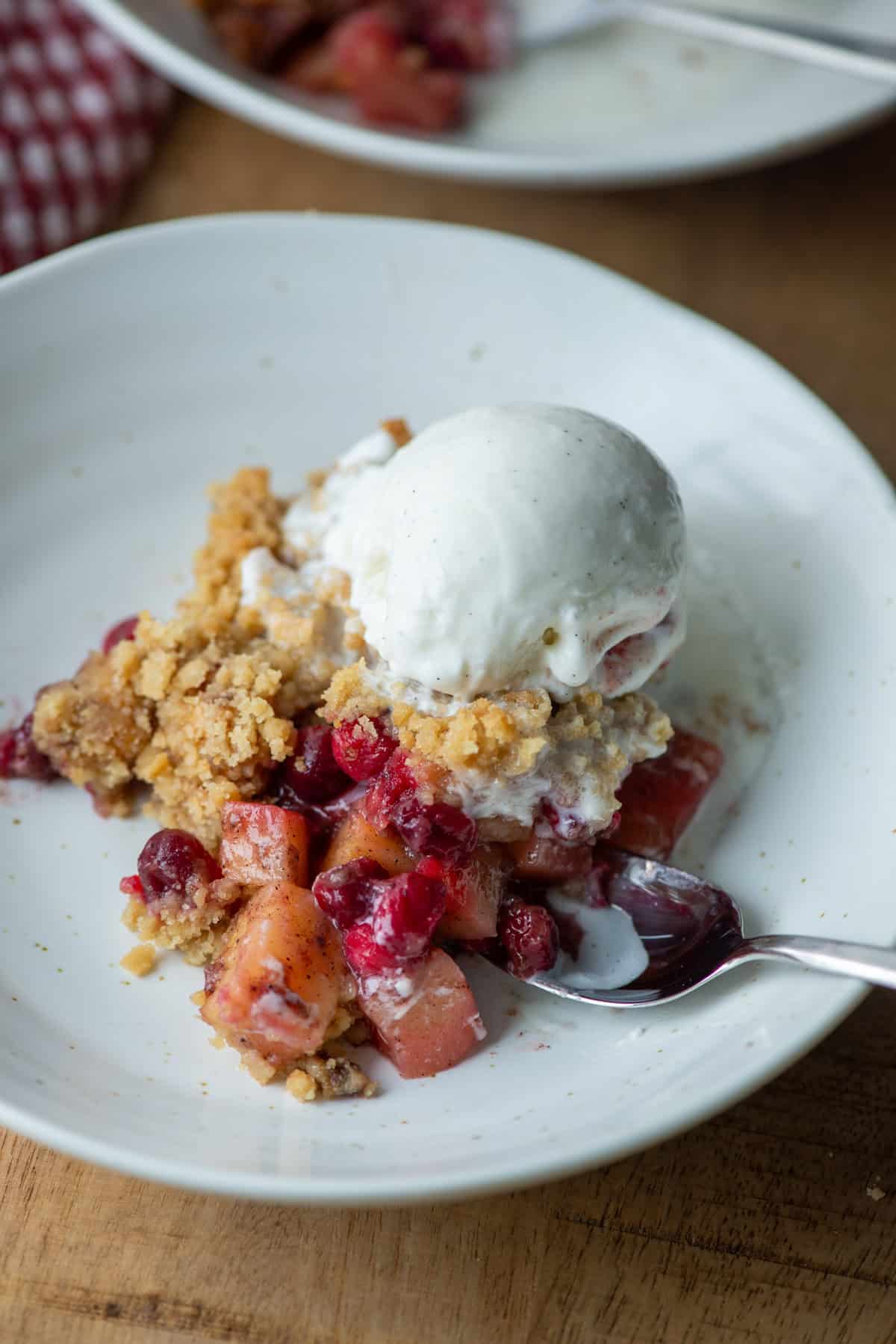 Close up of cranberry apple crumple on a plate with ice cream melting on top and a spoon sticking out of the side with another plate with cranberry apple crumble in the background
