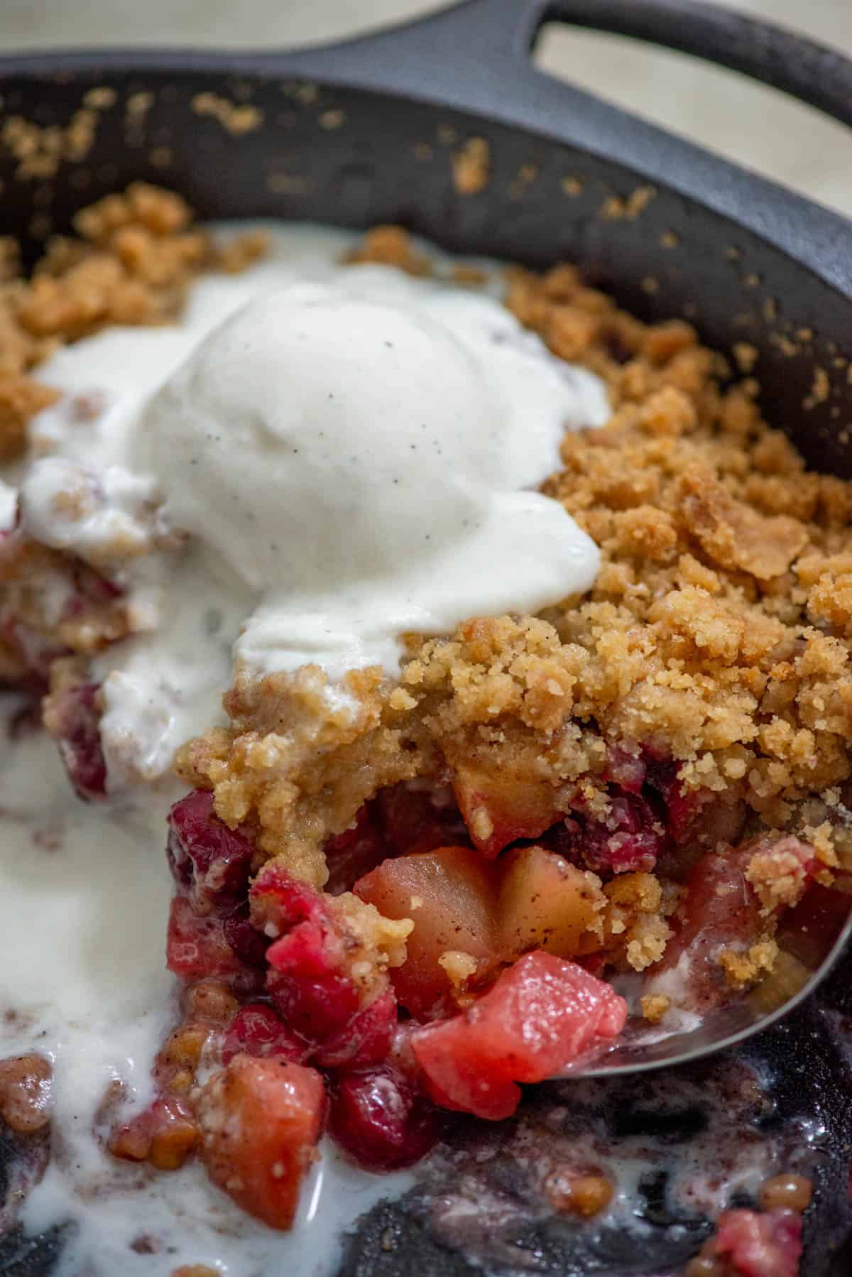 Overhead angled photo of cranberry apple crumble in a cast iron pan with a scoop of vanilla bean ice cream melting on top