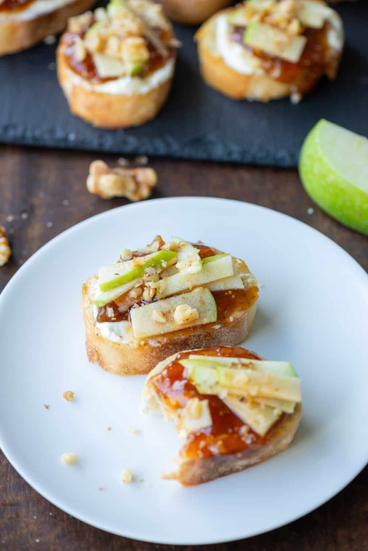 Overhead view of two apple, goat cheese and fig jam crostini on an appetizer plate in front to a platter of more crostini