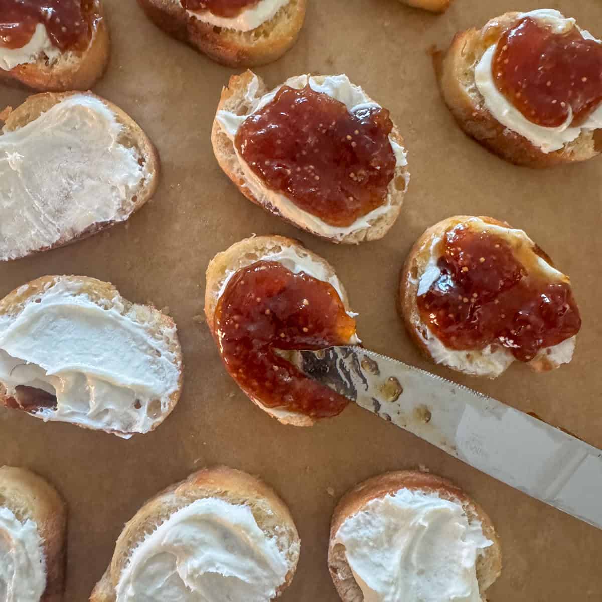 Overhead photo showing toasted baguette with whipped goat cheese where fig jam is being spread on top of the goat cheese