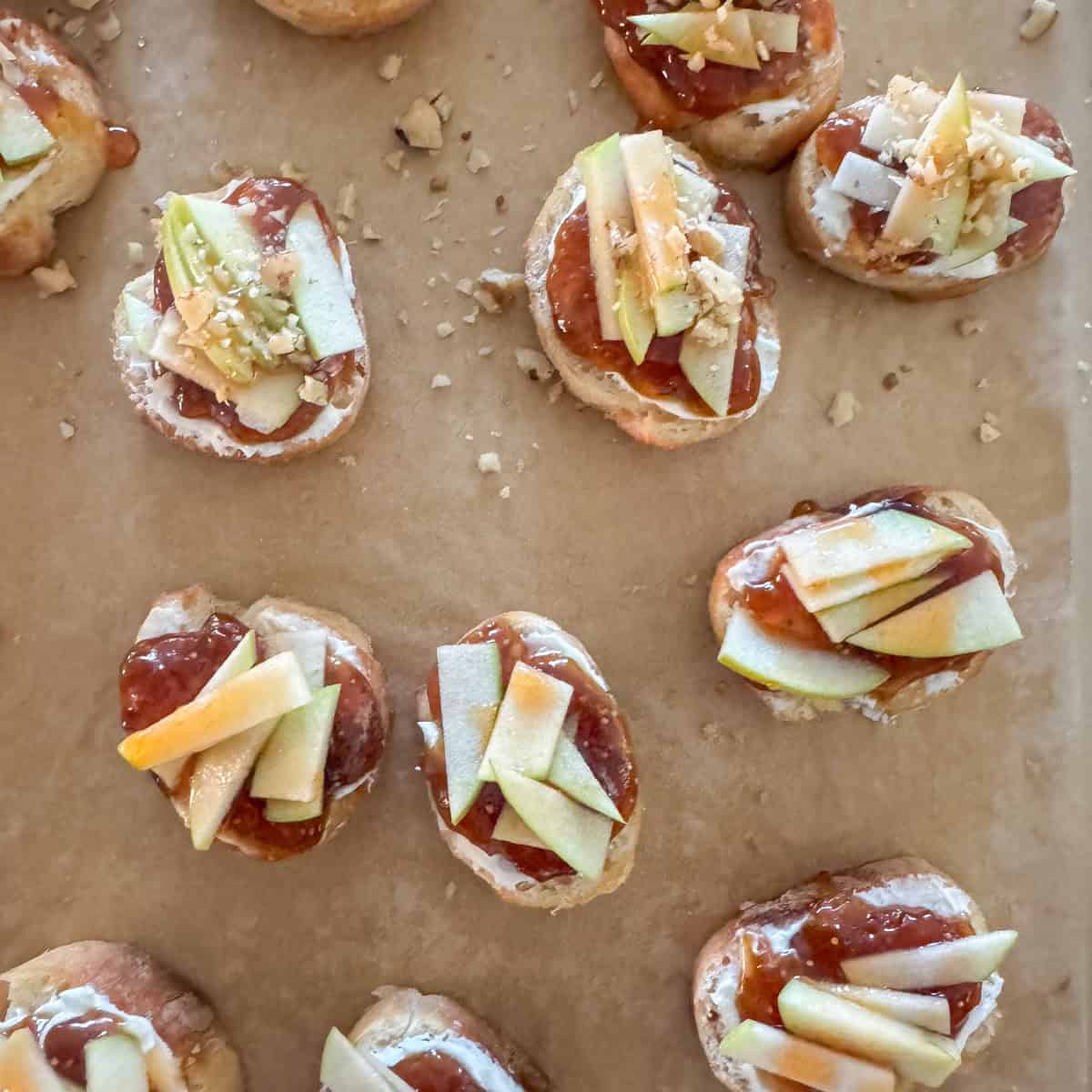 Overhead photo of last stage of making apple, goat cheese and fig jam crostini where sliced apple is place on top of the fig jam and then the crostini are toped with a drizzle of hot honey and chopped walnuts