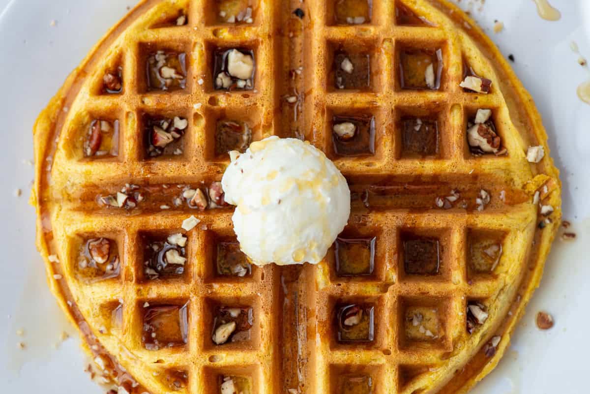 Overhead view of a pumpkin spice waffle topped with maple syrup, chopped pecans and whipped butter on a plate.