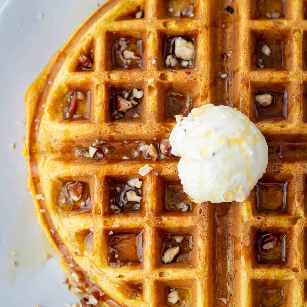 Overhead partial view of a pumpkin spice waffle topped with maple syrup, chopped pecans and whipped butter on a plate.