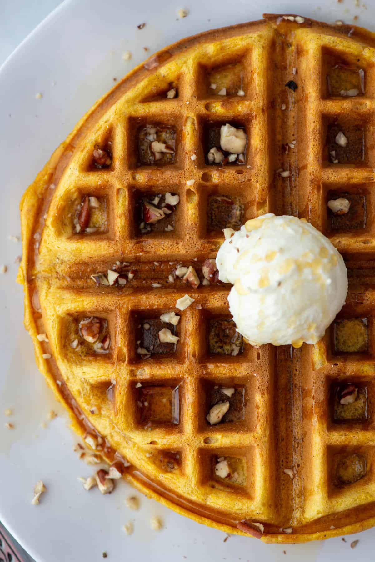 Overhead view of a pumpkin spice waffle topped with maple syrup, chopped pecans and whipped butter on a plate.